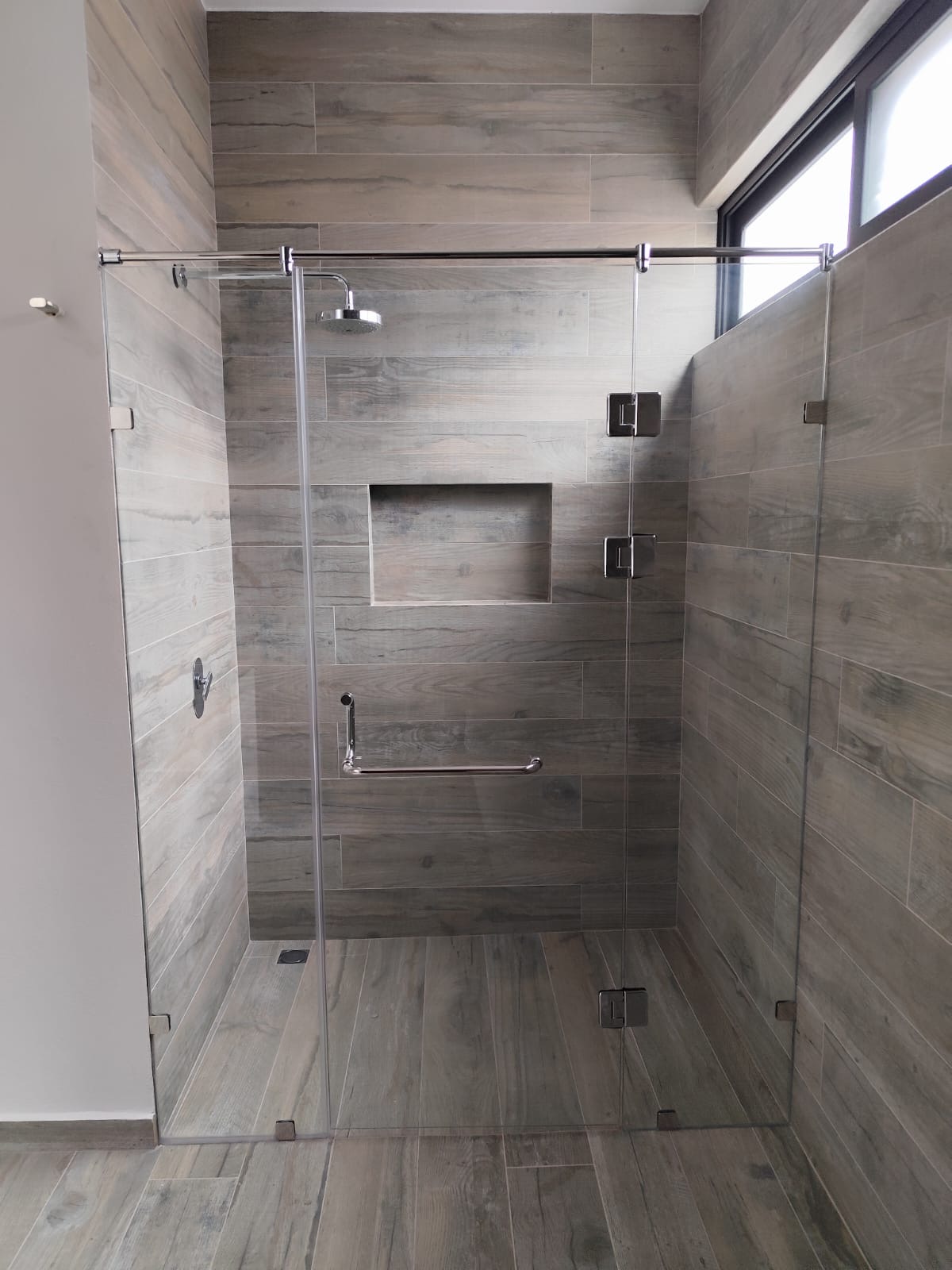 Cancel de baño esquinero de vidrio templado con herrajes cromados sobre muro de porcelanato imitación madera